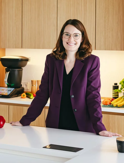 Vorwerk: Foto: Auf diesem Bild blickt Ramona Wehling direkt in die Kamera und stützt sich auf der Küchenarbeitsfläche ab. Sie guckt sympathisch in die Kamera. Hinter ihr steht der neue Thermomix® TM7.