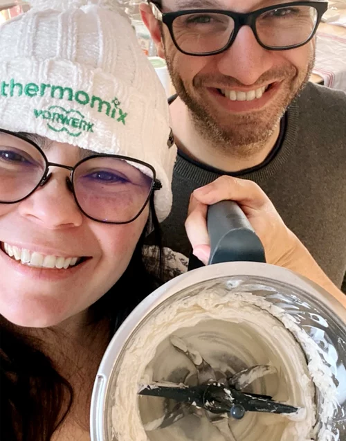 Vorwerk: Photo: A cheerful couple laughing and holding a Thermomix® bowl full of dough up to the camera. The woman is wearing a hat with the Thermomix® logo. Both look very happy.