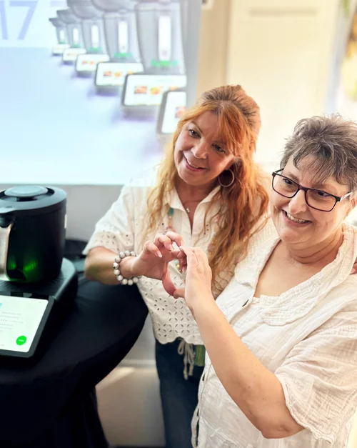 Vorwerk: Photo: Two women in work clothes, as is customary in care facilities, stand in a modern kitchen and give a thumbs-up. Next to them is a Thermomix® TM6, which they are obviously happy about.