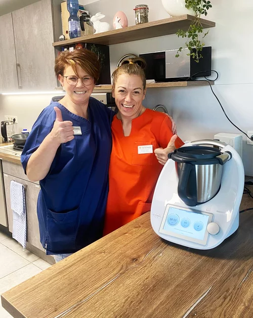 Vorwerk: Photo: Two middle-aged women standing close together in a kitchen, smiling and forming the heart sign with one hand each. Two friends. Next to them is the brand new Thermomix® TM7.