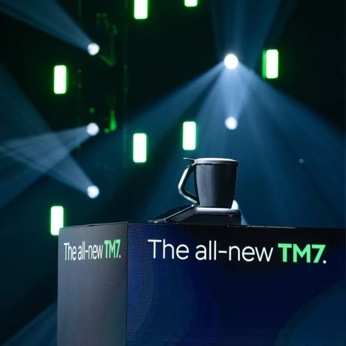 Vorwerk: Foto: Das Gerät allein auf der Bühne, schwarz vor schwarzem Hintergrund, der von grünen und blauen Lichtpunkten erhellt ist. Auf dem kleinen Podest, auf dem der TM7 steht, ist zu lesen: „the all-new TM7“. Zu deutsch: Der völlig neue TM7.
