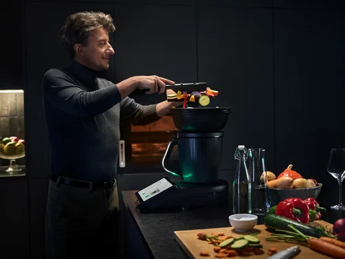 Vorwerk: Foto: Ein Mann, von der Seite fotografiert, füllt in einer ganz in Schwarz eingerichteten Küche den neuen TM7 mit allerlei Gemüse. Vor ihm auf der Arbeitsfläche liegt noch mehr frischgeschnittenes Gemüse wie Möhren, Zucchini, Paprika und Zwiebeln.