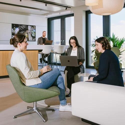 Vorwerk: Foto: Dieses Foto zeigt einen der offen gestalteten Meeting points, zu deutsch: Treffpunkte, im neuen, zeitgemäßen Büroareal. Im Vordergrund sitzen drei Kolleginnen auf Sesseln und Sofa. Im Hintergrund bedient sich ein Kollege an der Getränkebar.