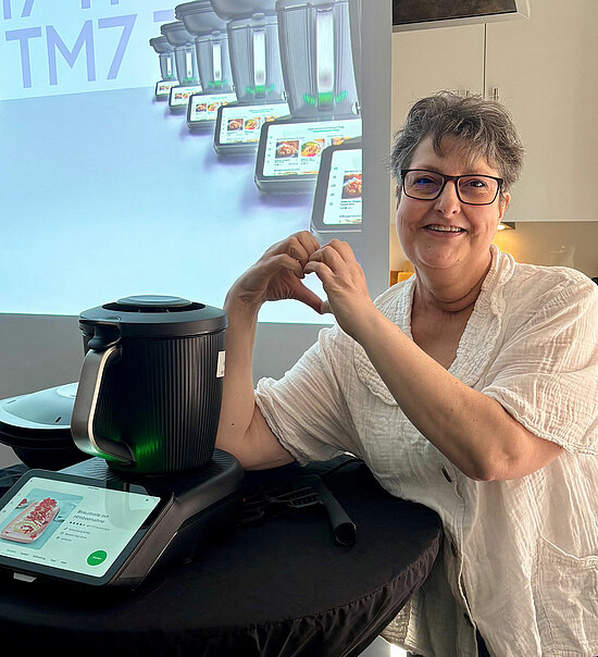 Vorwerk: Photo: An elderly woman forms a heart with her hands. A TM7 can be seen next to her.