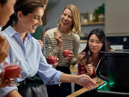 Vorwerk: Foto: Vier Frauen stehen vor dem grün leuchtenden Thermomix in einer Küche und strahlen übers ganz Gesicht. In den Händen halten sie Dessertgläser und -löffel mit einem roten Nachtisch darin.
