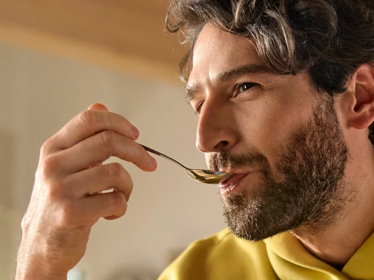 Vorwerk: Foto: Ein Mann mit dichten Locken und Dreitage-Bart hat einen Löffel im Mund, um etwas zu probieren, und macht ein genüssliches Gesicht.