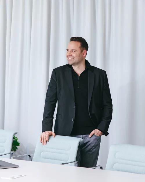 Vorwerk: Foto: Julius Ganns, bei Vorwerk für Software und Digitales verantwortlich, steht entspannt an einem Schreibtisch, blickt lächelnd nach rechts in die Ferne.